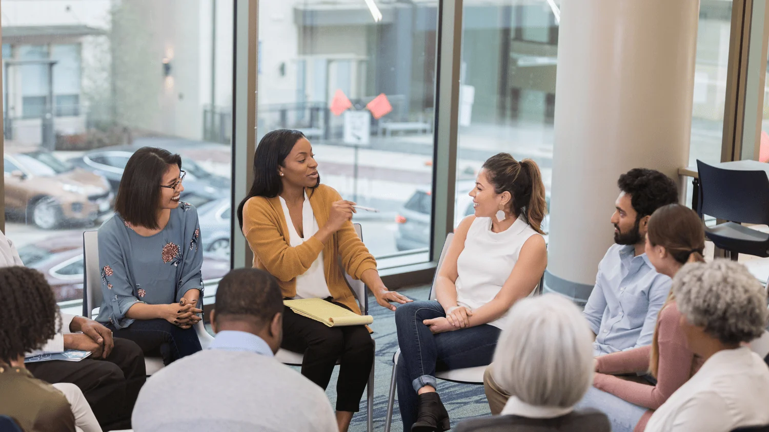 A facilitator leads a seated group discussion in a bright, sunlit meeting space