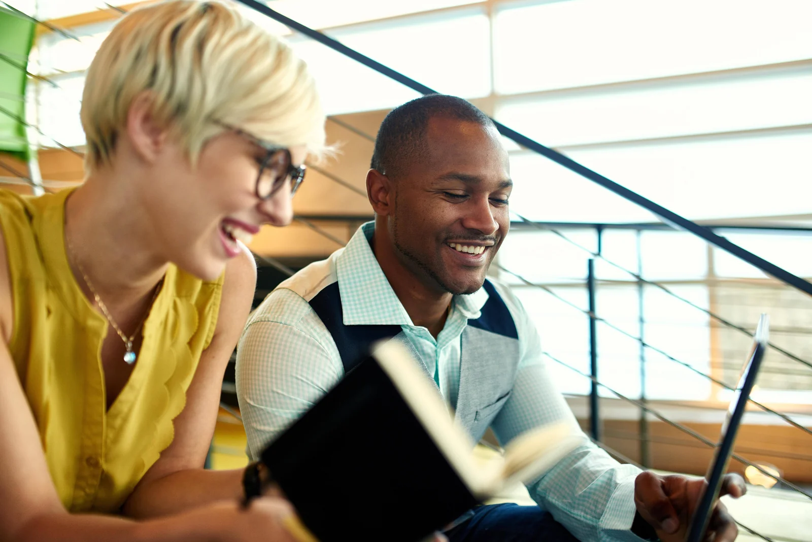 Two colleagues laughing together over a notebook on an office staircase