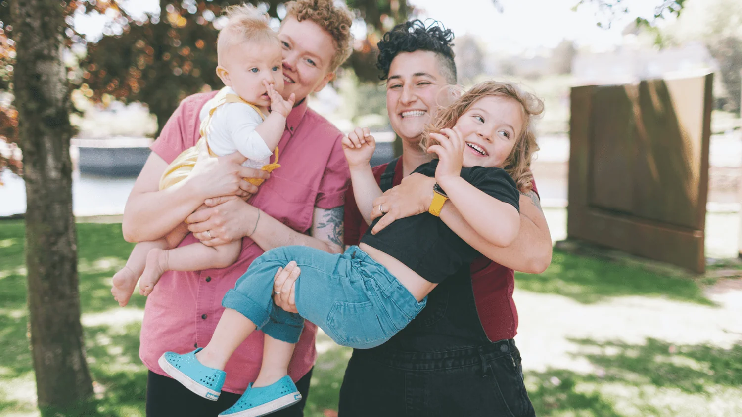 Two parents laughing outdoors in a park while holding their baby and young child