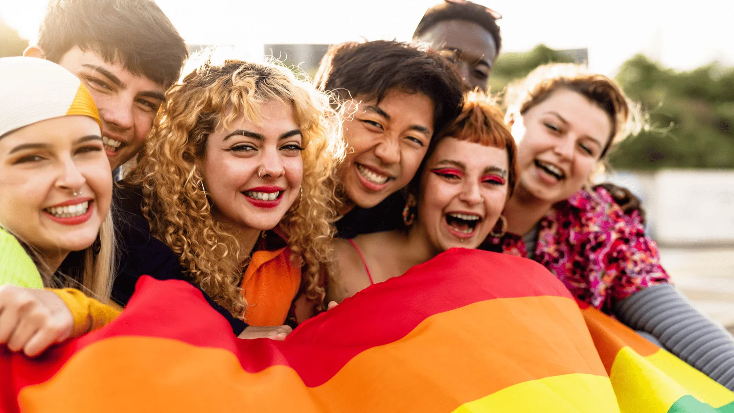 A group of young people laughing together while holding a rainbow Pride flag in the sunshine
